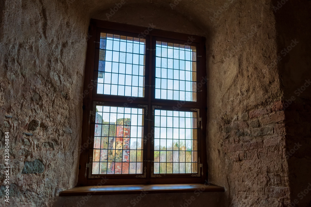 Autumn day light going through vintage stained-glass window in very thick wall niche in the dark medieval castle. Ruins of medieval castle behind the window.