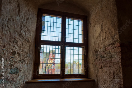Autumn day light going through vintage stained-glass window in very thick wall niche in the dark medieval castle. Ruins of medieval castle behind the window.