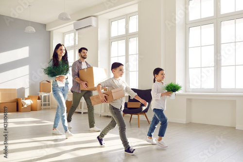 Moving day and relocation concept. Happy excited caucasian family with children carrying cardboard boxes and green houseplants to new home. Bright apartment house interior with lack of furniture