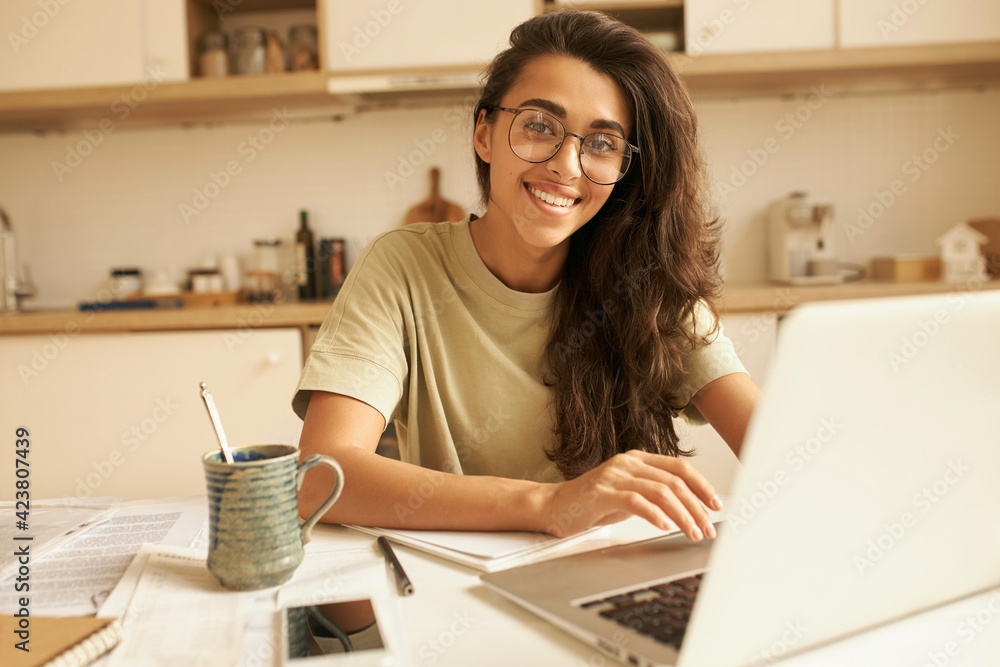 Successful young businesswoman sitting in kitchen having coffee, using laptop for remote work, checking e-mail, communicating with customers online. Cute student girl in eyeglasses doing homework Successful young businesswoman sitting in kitchen having coffee, using laptop for remote work, checking e-mail, communicating with customers online. Cute student girl in eyeglasses doing homework