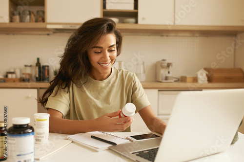 Nutrition, healthy lifestyle and body hacking. Joyful young woman nutritionist sitting in front of open laptop, having online consultation, entering information about dietary supplement in her hand photo