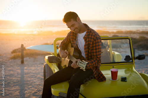 Fototapeta Happy caucasian man sitting on beach buggy by the sea playing guitar during sunset