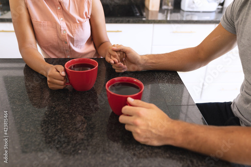 Diverse couple sitting in kitchen drinking coffee and holding hands