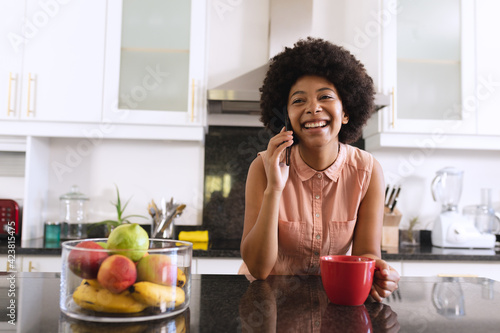 African american woman in kitchen drinking coffee and talking on smartphone