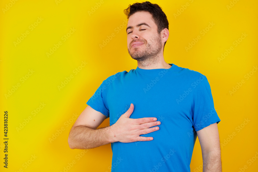 young handsome caucasian man wearing blue t-shirt against yellow background touches tummy, smiles gently, eating and satisfaction concept.