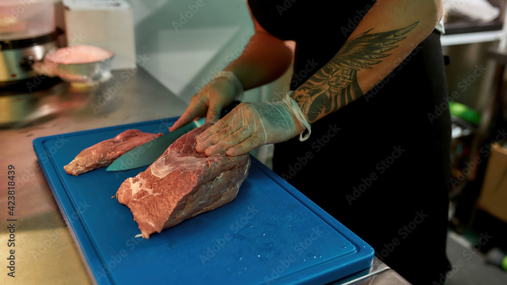 Close up shot of male cook wearing protective gloves slicing raw pork meat on a plastic board while working in commercial kitchen