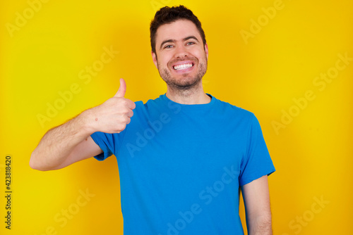 young handsome caucasian man wearing blue t-shirt against yellow background doing happy thumbs up gesture with hand. Approving expression looking at the camera showing success.