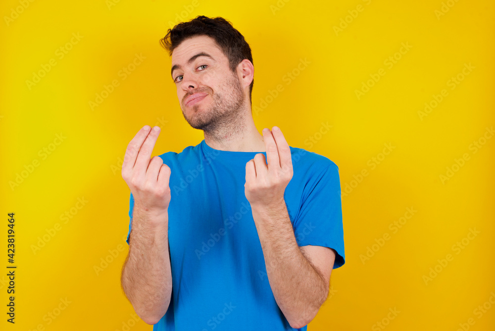 young handsome caucasian man wearing blue t-shirt against yellow background doing money gesture with hands, asking for salary payment, millionaire business young handsome caucasian man wearing blue t-shirt against yellow background doing money gesture with hands, asking for salary payment, millionaire business