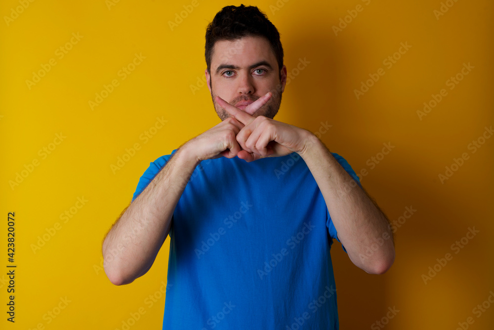 young handsome caucasian man wearing blue t-shirt against yellow background Has rejection angry expression crossing fingers doing negative sign. young handsome caucasian man wearing blue t-shirt against yellow background Has rejection angry expression crossing fingers doing negative sign.