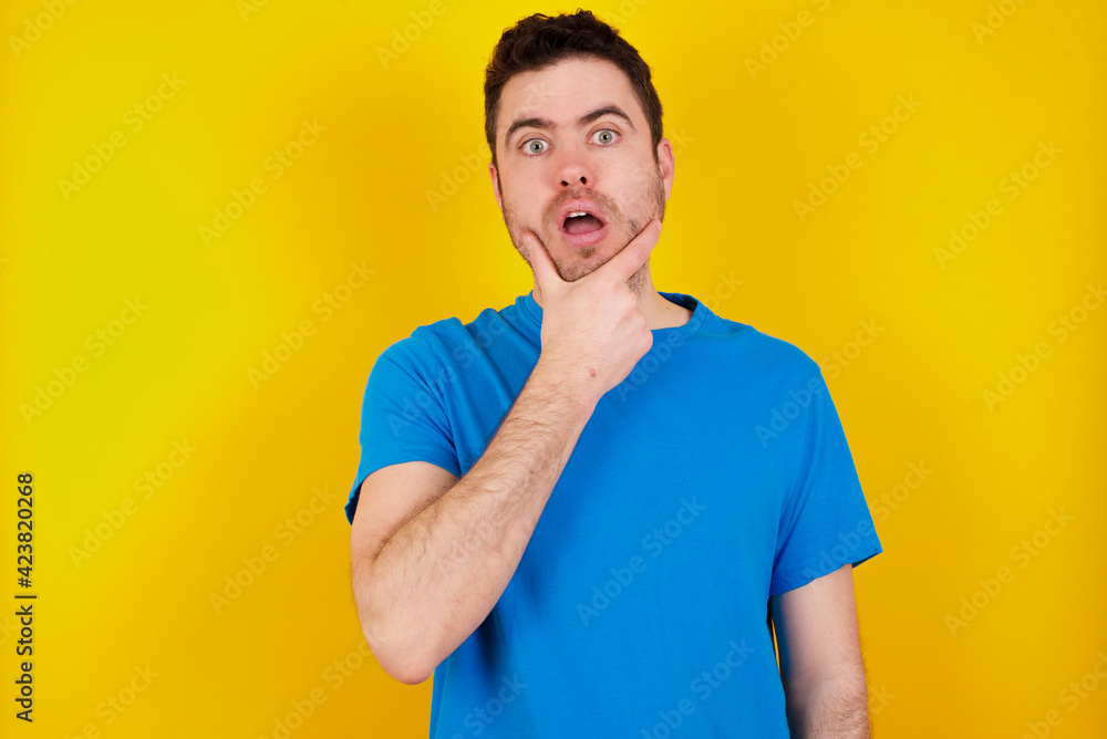 young handsome caucasian man wearing blue t-shirt against yellow background Looking fascinated with disbelief, surprise and amazed expression with hands on chin young handsome caucasian man wearing blue t-shirt against yellow background Looking fascinated with disbelief, surprise and amazed expression with hands on chin