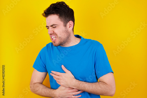 young handsome caucasian man wearing blue t-shirt against yellow background with hand on stomach because nausea, painful disease feeling unwell. Ache concept.
