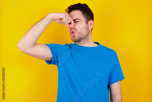 young handsome caucasian man wearing blue t-shirt against yellow background smelling something stinky and disgusting, intolerable smell, holding breath with fingers on nose. Bad smell