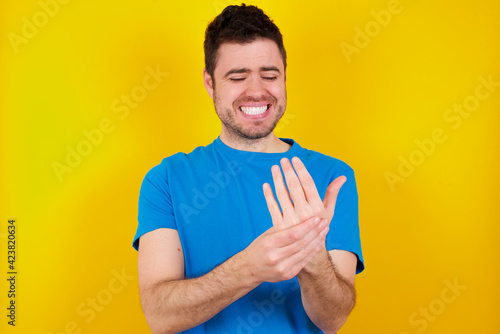 young handsome caucasian man wearing blue t-shirt against yellow background asking to be quiet with finger on lips pointing with hand to the side. Silence and secret concept.