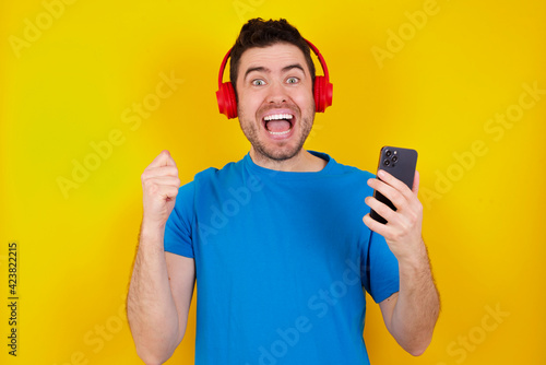 Happy young handsome caucasian man wearing blue t-shirt against yellow background feels good while focused in screen of smartphone. People, technology, lifestyle