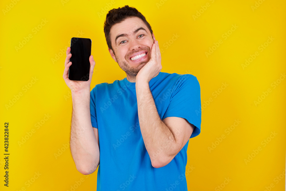 Smiling young handsome caucasian man wearing blue t-shirt against yellow background Mock up copy space. Pointing index finger on mobile phone with blank empty screen Smiling young handsome caucasian man wearing blue t-shirt against yellow background Mock up copy space. Pointing index finger on mobile phone with blank empty screen