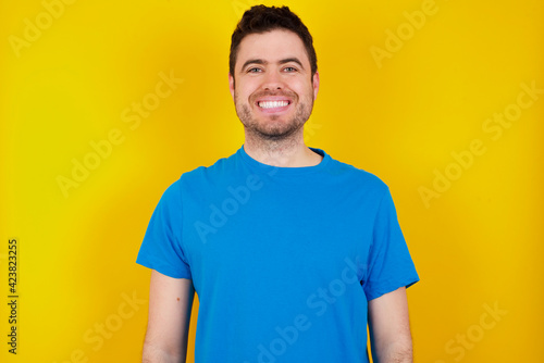 young handsome caucasian man wearing blue t-shirt against yellow background holding an invisible aligner and pointing at it. Dental healthcare and confidence concept.