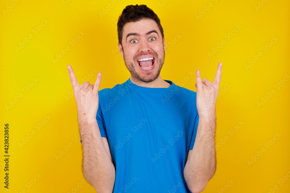 young handsome caucasian man wearing blue t-shirt against yellow background makes rock n roll sign looks self confident and cheerful enjoys cool music at party. Body language concept. young handsome caucasian man wearing blue t-shirt against yellow background makes rock n roll sign looks self confident and cheerful enjoys cool music at party. Body language concept.