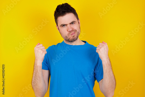 Irritated young handsome caucasian man wearing blue t-shirt against yellow background blows cheeks with anger and raises clenched fists expresses rage and aggressive emotions. Furious model