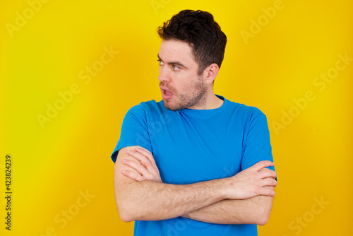 Astonished young handsome caucasian man wearing blue t-shirt against yellow background looks aside surprisingly with opened mouth.