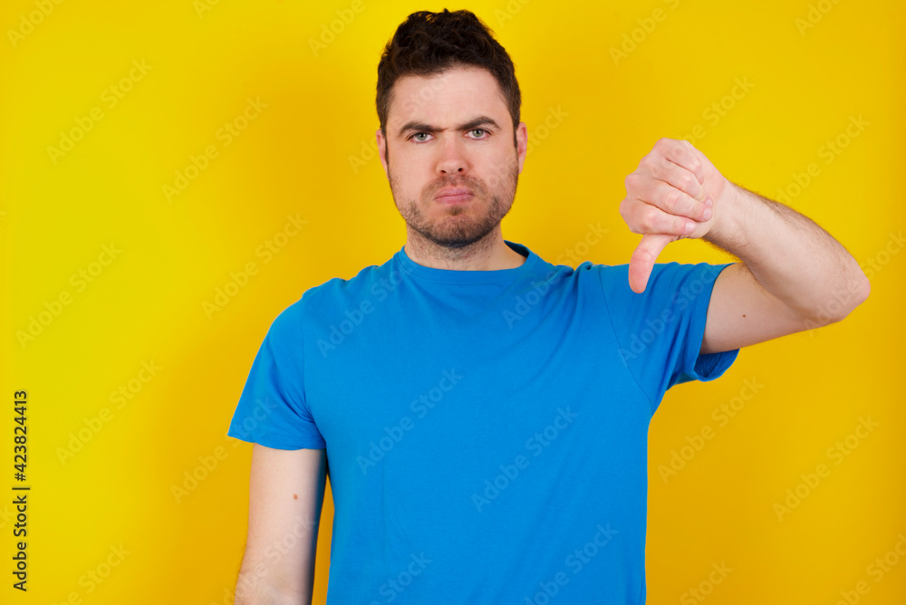 young handsome caucasian man wearing blue t-shirt against yellow background looking unhappy and angry showing rejection and negative with thumbs down gesture. Bad expression. young handsome caucasian man wearing blue t-shirt against yellow background looking unhappy and angry showing rejection and negative with thumbs down gesture. Bad expression.