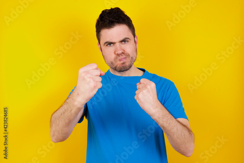 Displeased annoyed young handsome caucasian man wearing blue t-shirt against yellclenches fists, gestures pissed, ready to revenge, looks with aggression at camera stands full of hate, being pressured