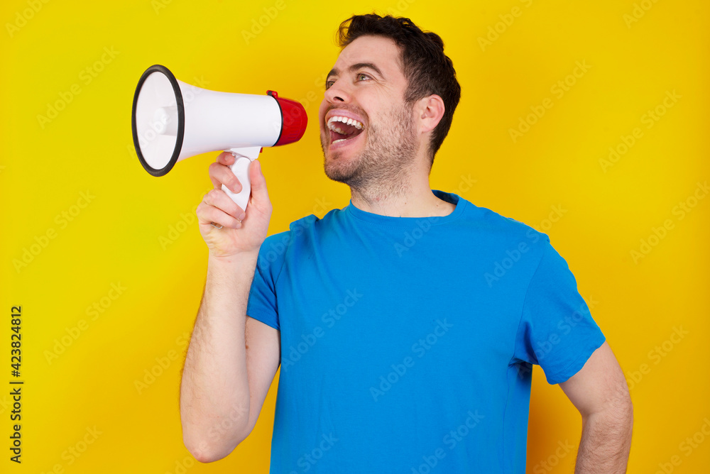 Funny young handsome caucasian man wearing blue t-shirt against yellow background People sincere emotions lifestyle concept. Mock up copy space. Screaming in megaphone. Funny young handsome caucasian man wearing blue t-shirt against yellow background People sincere emotions lifestyle concept. Mock up copy space. Screaming in megaphone.