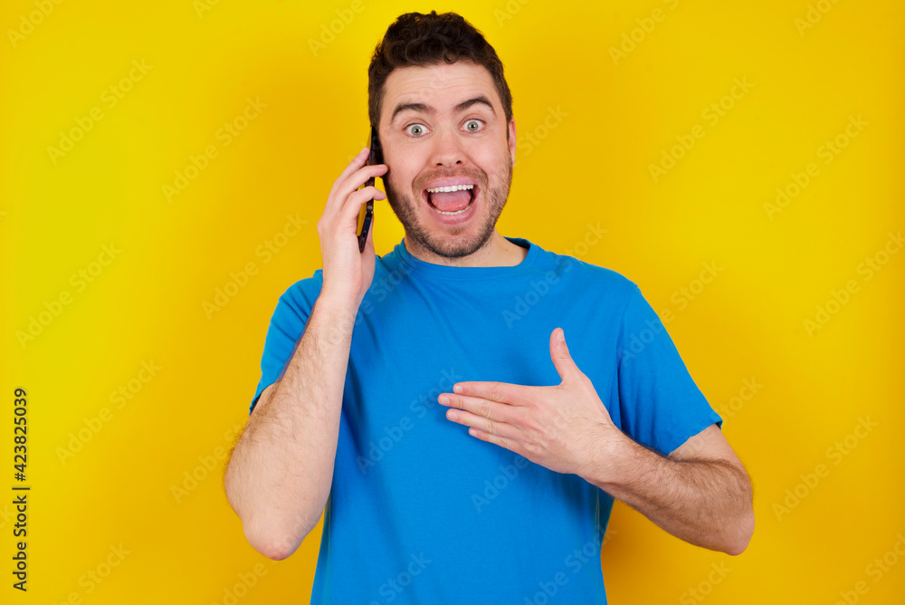 Smiling young handsome caucasian man wearing blue t-shirt against yellow background talks via cellphone, enjoys pleasant great conversation. People, technology, communication concept Smiling young handsome caucasian man wearing blue t-shirt against yellow background talks via cellphone, enjoys pleasant great conversation. People, technology, communication concept