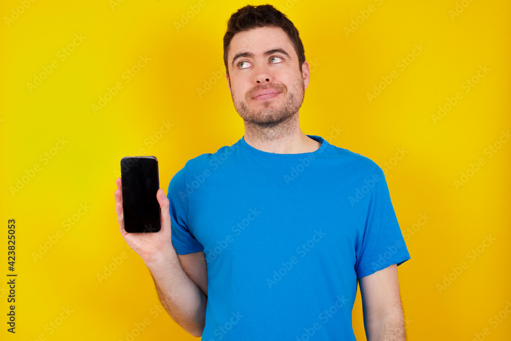 young handsome caucasian man wearing blue t-shirt against yellow background holds new mobile phone and looks mysterious aside shows blank display of modern cellular young handsome caucasian man wearing blue t-shirt against yellow background holds new mobile phone and looks mysterious aside shows blank display of modern cellular