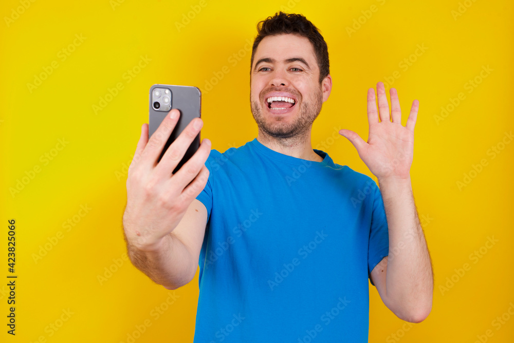 young handsome caucasian man wearing blue t-shirt against yellow background holds modern mobile phone and makes video call waves palm in hello gesture. People modern technology concept young handsome caucasian man wearing blue t-shirt against yellow background holds modern mobile phone and makes video call waves palm in hello gesture. People modern technology concept