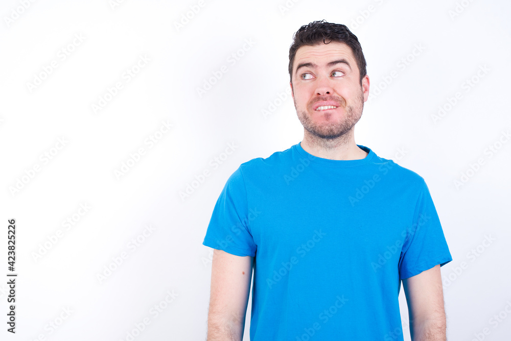 Amazed young handsome caucasian man wearing blue t-shirt against white background bitting lip and looking tricky to empty space. Amazed young handsome caucasian man wearing blue t-shirt against white background bitting lip and looking tricky to empty space.