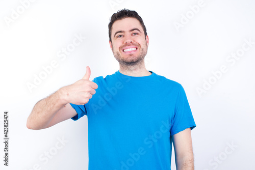 young handsome caucasian man wearing blue t-shirt against white background doing happy thumbs up gesture with hand. Approving expression looking at the camera showing success.
