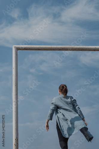 Back view of fierce confident walk of a woman dressed in an overside aqua cardigan with a blue sky background  framed by a football gate