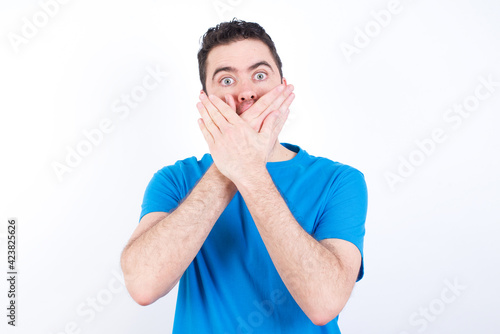 young handsome caucasian man wearing blue t-shirt against white background shocked covering mouth with hands for mistake. Secret concept.