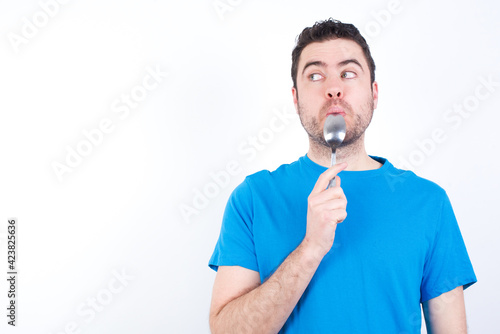 Very hungry young handsome caucasian man wearing blue t-shirt against white background holding spoon into mouth dream of tasty meal