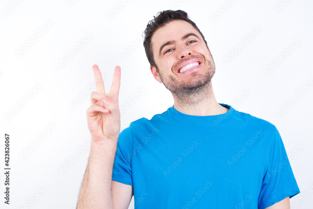 young handsome caucasian man wearing blue t-shirt against white background smiling with happy face winking at the camera doing victory sign. Number two. young handsome caucasian man wearing blue t-shirt against white background smiling with happy face winking at the camera doing victory sign. Number two.