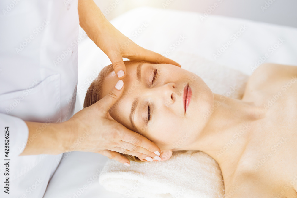 Beautiful caucasian woman enjoying facial massage with closed eyes in sunny spa salon. Relaxing treatment in medicine Beautiful caucasian woman enjoying facial massage with closed eyes in sunny spa salon. Relaxing treatment in medicine