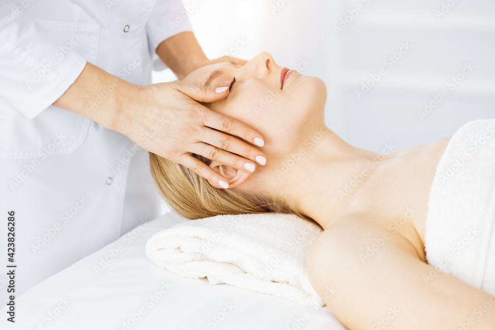 Beautiful caucasian woman enjoying facial massage with closed eyes in sunny spa salon. Relaxing treatment in medicine Beautiful caucasian woman enjoying facial massage with closed eyes in sunny spa salon. Relaxing treatment in medicine
