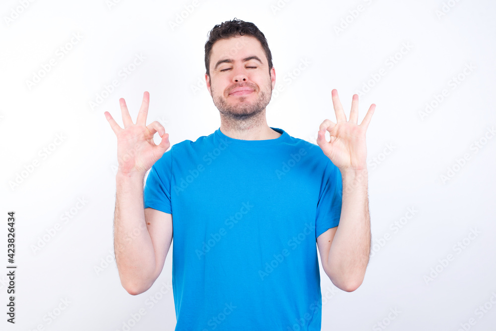 young handsome caucasian man wearing blue t-shirt against white background relax and smiling with eyes closed doing meditation gesture with fingers. Yoga concept. young handsome caucasian man wearing blue t-shirt against white background relax and smiling with eyes closed doing meditation gesture with fingers. Yoga concept.