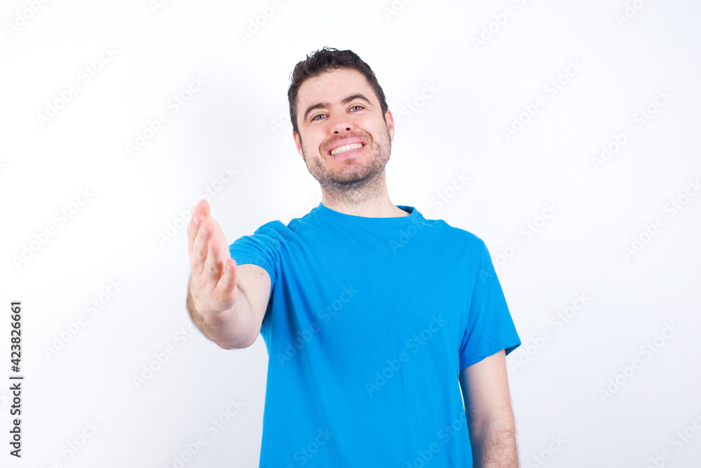 young handsome caucasian man wearing blue t-shirt against white background smiling friendly offering handshake as greeting and welcoming. Successful business. young handsome caucasian man wearing blue t-shirt against white background smiling friendly offering handshake as greeting and welcoming. Successful business.