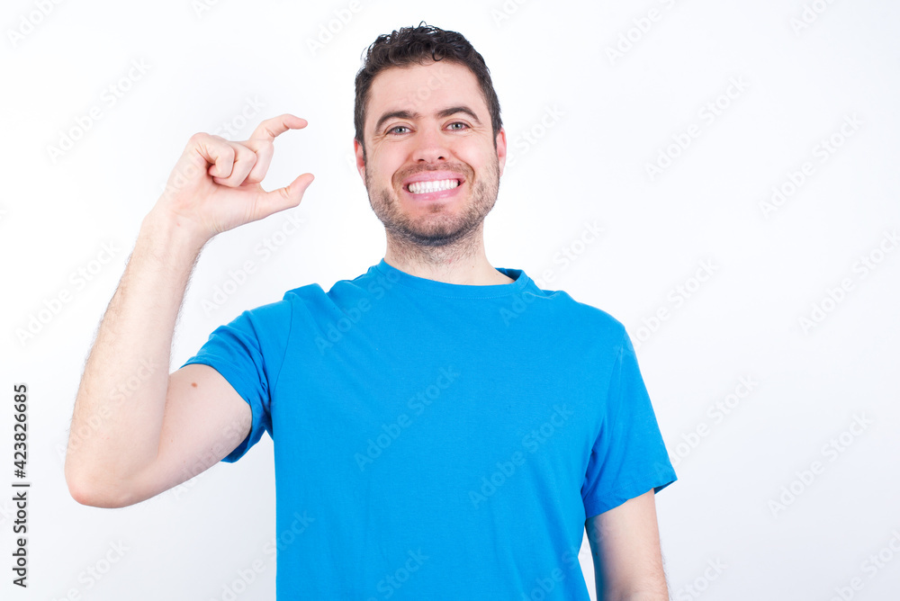 young handsome caucasian man wearing blue t-shirt against white background smiling and confident gesturing with hand doing small size sign with fingers looking and the camera. Measure concept young handsome caucasian man wearing blue t-shirt against white background smiling and confident gesturing with hand doing small size sign with fingers looking and the camera. Measure concept