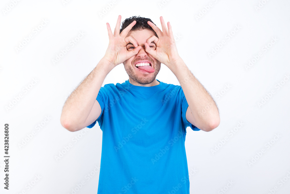young handsome caucasian man wearing blue t-shirt against white background doing ok gesture like binoculars sticking tongue out, eyes looking through fingers. Crazy expression. young handsome caucasian man wearing blue t-shirt against white background doing ok gesture like binoculars sticking tongue out, eyes looking through fingers. Crazy expression.