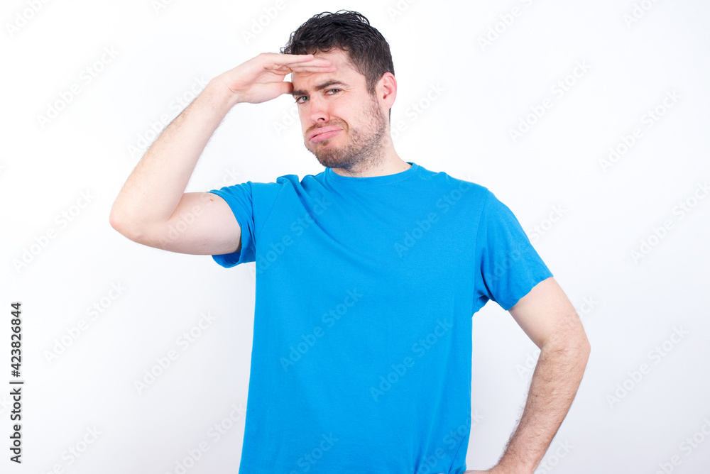 young handsome caucasian man wearing blue t-shirt against white background having problems, worried and stressed holds hand on forehead. young handsome caucasian man wearing blue t-shirt against white background having problems, worried and stressed holds hand on forehead.