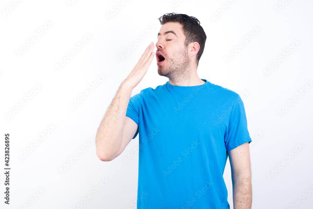 young handsome caucasian man wearing blue t-shirt against white background being tired and yawning after spending all day at work. young handsome caucasian man wearing blue t-shirt against white background being tired and yawning after spending all day at work.