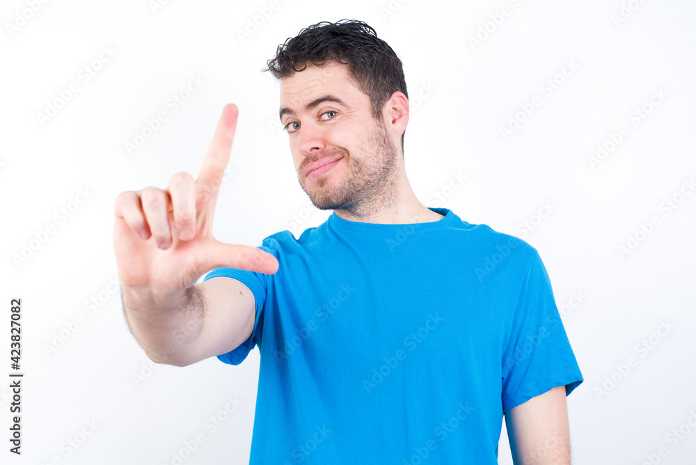 young handsome caucasian man wearing blue t-shirt against white background making fun of people with fingers on forehead doing loser gesture mocking and insulting. young handsome caucasian man wearing blue t-shirt against white background making fun of people with fingers on forehead doing loser gesture mocking and insulting.