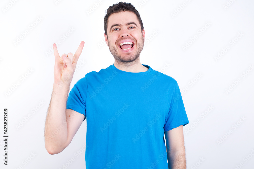young handsome caucasian man wearing blue t-shirt against white background doing a rock gesture and smiling to the camera. Ready to go to her favorite band concert. young handsome caucasian man wearing blue t-shirt against white background doing a rock gesture and smiling to the camera. Ready to go to her favorite band concert.