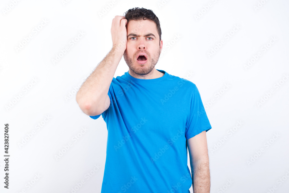 Embarrassed young handsome caucasian man wearing blue t-shirt against white background with shocked expression, expresses great amazement, Puzzled model poses indoor Embarrassed young handsome caucasian man wearing blue t-shirt against white background with shocked expression, expresses great amazement, Puzzled model poses indoor