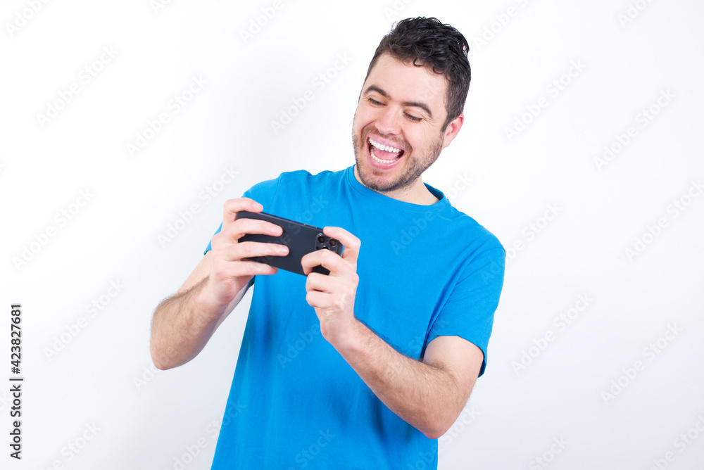 young handsome caucasian man wearing blue t-shirt against white background holding in hands cell playing video games or chatting young handsome caucasian man wearing blue t-shirt against white background holding in hands cell playing video games or chatting