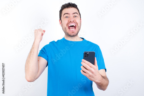 Portrait of young handsome caucasian man wearing blue t-shirt against white background holding in hands cell celebrating