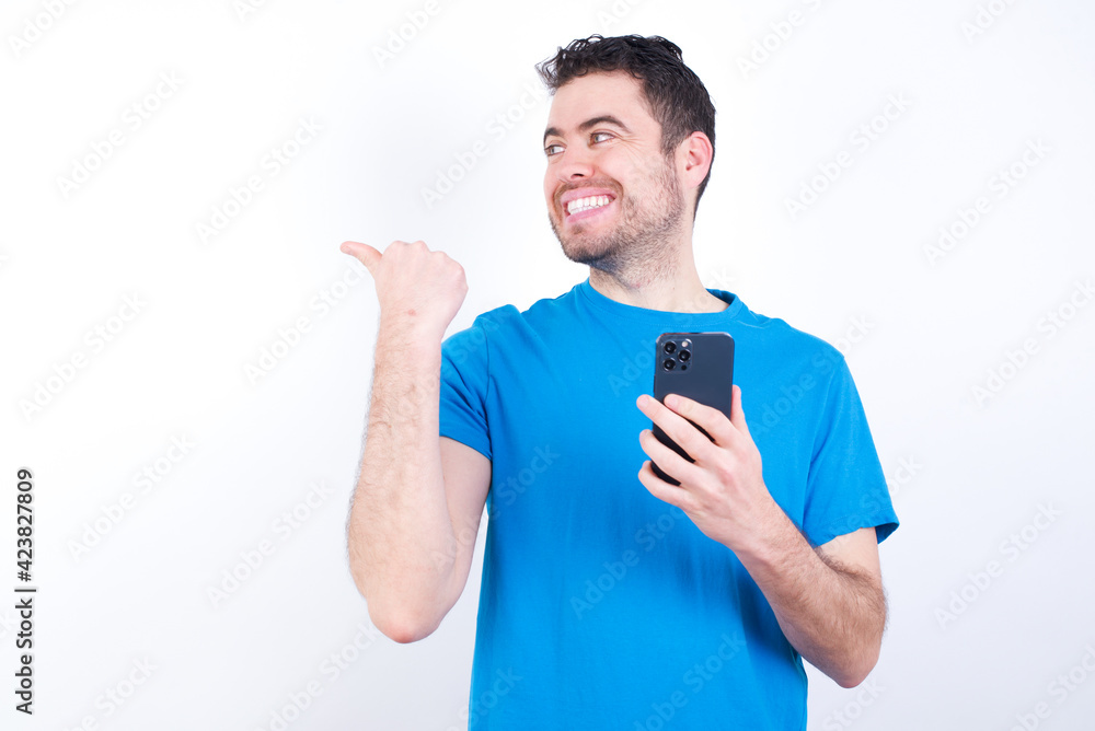 young handsome caucasian man wearing blue t-shirt against white background using and texting with smartphone pointing and showing with thumb up to the side with happy face smiling young handsome caucasian man wearing blue t-shirt against white background using and texting with smartphone pointing and showing with thumb up to the side with happy face smiling
