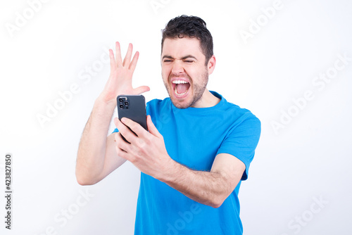 Angry young handsome caucasian man wearing blue t-shirt against white background screaming on the phone, having an argument with an employee. Troubles at work.